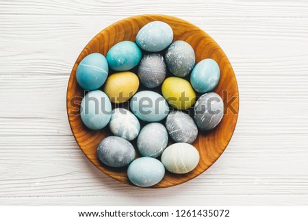 Stylish easter eggs in wooden plate on white wooden background, flat lay. Modern easter eggs painted with natural dye in blue, grey, yellow marble color. Happy Easter