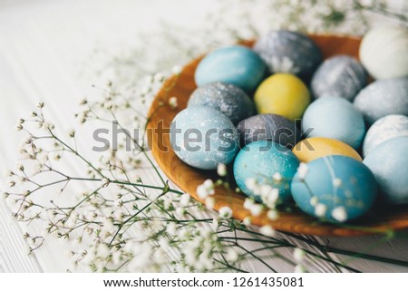 Stylish easter eggs with spring flowers on wooden plate on white wooden background. Modern easter eggs painted with natural dye in blue, grey, yellow marble color. Happy Easter