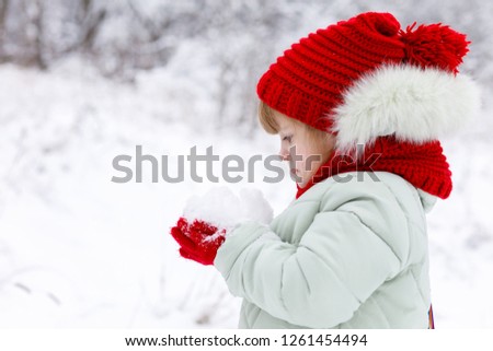 A little cute girl in bright clothes with funny emotions on her face is standing sidelong and holding a heap of snow in her hand at winter park background.