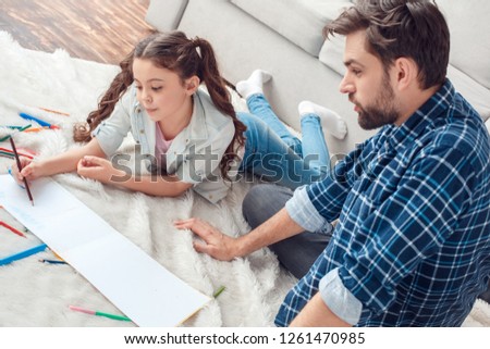 Bearded man and little girl at home family time on carpet on floor father looking at daughter drawing picture with color pencils concentrated