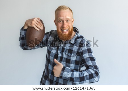 Red hair mature man standing isolated on grey wall holding american football ball looking camera smiling cheerful showing thumb up close-up