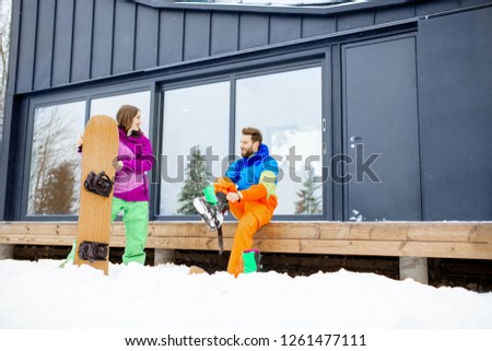 Young couple in ski suits preparing for snowboarding sitting on the terrace of the modern house in the mountains