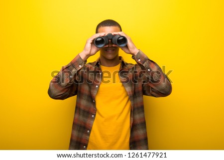 Young african american man on vibrant yellow background and looking for something in the distance with binoculars