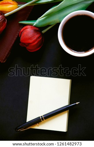 beautiful top view close up photo of blank notepad with copy space,pencil,red tulips and white cup of coffee on dark black background 