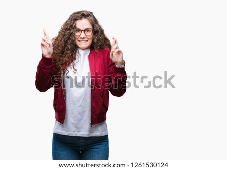 Beautiful brunette curly hair young girl wearing jacket and glasses over isolated background smiling crossing fingers with hope and eyes closed. Luck and superstitious concept.