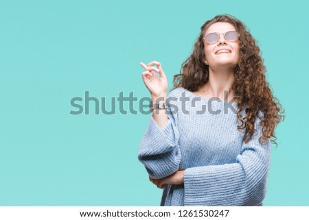 Beautiful brunette curly hair young girl wearing sunglasses over isolated background with a big smile on face, pointing with hand and finger to the side looking at the camera.