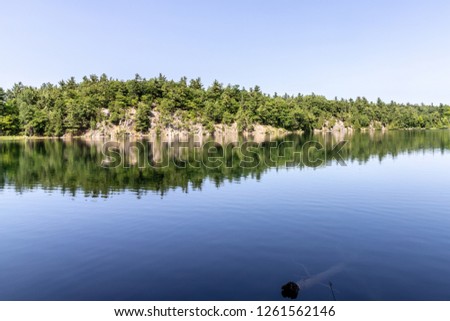 Horizontal landscape of trees and a rocky hill shoreline with beautiful reflections in the clear water of Pink Lake on a sunny day in Gatineau Park, Quebec