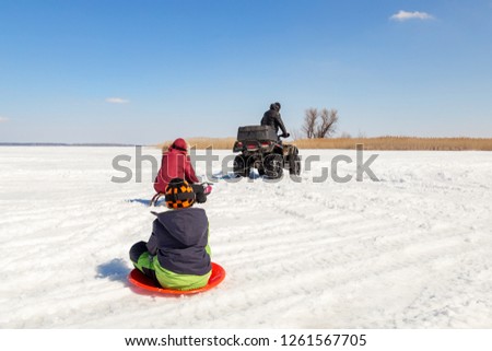 Man on ATV quadbike riding sledges with kids in tow on frozen lake surface at winter. Winter extreme sports and recreation. Children outdoor fun and activities