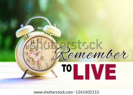 A retro alarm clock on a white table top with nature background and morning sunlight. A text 'Remember to live' written on image.