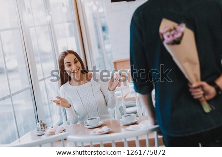 Couple in Cafe. Couple is Beautiful Young Man and Woman. Man is Holding Bouquet of Flowers Behind His Back. Flowers is Surprise For Girl. Girl is Surprised. Persons is Sits at Table. Sunny Daytime.