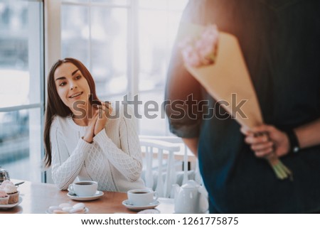 Couple Resting in Cafe. Couple is Beautiful Young Man and Woman. Man is Holding Bouquet of Flowers Behind His Back. Flowers is Surprise For Girl. Persons is Sitting at Table. Sunny Daytime.