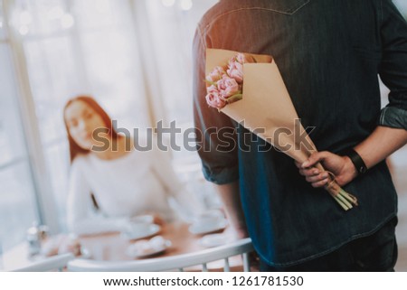 Couple in Cafe. Couple is Young Man and Woman. Man is Holding Bouquet of Flowers Behind His Back. Flowers is Surprise For Girl. Blur Image of Girl. Persons is Sitting at Table. Sunny Daytime.