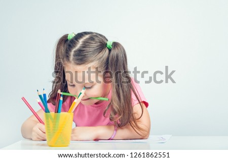 beautiful cute little girl holding pencil in mouth and drawing picture on white paper