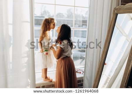 Young mother kisses her little daughter standing on the windowsill next to the mirror in the full of light cozy room