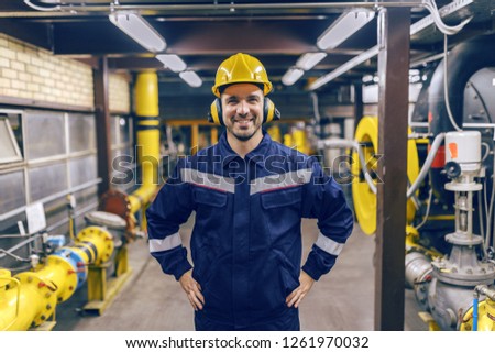 Young smiling Caucasian worker inn protective suit with hands on hips standing in heat plant. Royalty-Free Stock Photo #1261970032