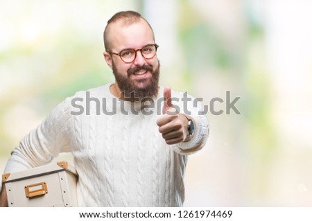 Young hipster man wearing glasses, moving holding moving box over isolated background happy with big smile doing ok sign, thumb up with fingers, excellent sign