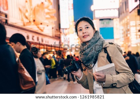 girl tourist smiling holding camera and guidebook standing on the street in dotonbori in osaka japan at night with dark sky in background. busy teeming japanese urban city lifestyle concept.