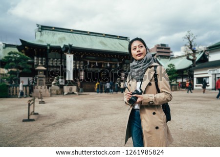 beautiful young asian woman holding slr camera sightseeing in historical architecture made by wood. lady photographer visiting tenmangu temple in osaka japan smiling standing on ground.