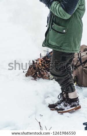 Girl is sitting near camp fire an in winter time. Concept adventure active vacations outdoor. Winter camping. Snowy forest. vintage backpack and thermos with tea, coffee, vertical photo