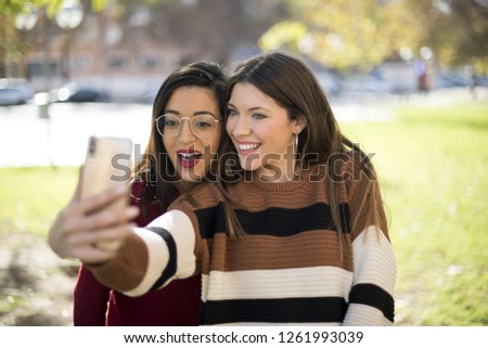 Multi ethnic friends taking a selfie standing outdoors. Two joyful cheerful girls taking a selfie while sitting together at the park having fun and smiling at the phone  - Imagen