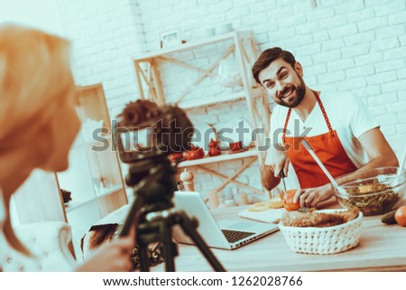Blogger Makes a Video. Blogger is Smiling Beard Man. Video About a Cooking. Woman Operator Shoots a Video on Camera. Different Food on Table. Man Cutting a Peppers. People in Studio Interior.