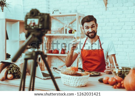 Blogger Makes a Video. Blogger is Smiling Beard Man. Video About a Cooking. Camera Shoots a Video. Laptop and Different Food on Table. Man Showing a White Wine in Wineglass. Man in Studio Interior.