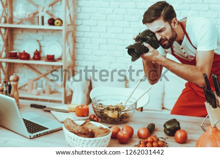 Photographer Makes a Photo. Photographer is Young Beard Man. Man is Taking Picture of Cooking a Food. Guy is Using a Photo Camera. Laptop and Different Food on Table. Man in Studio Interior.