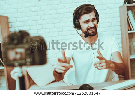 Blogger Makes a Video. Blogger is Gamer. Blogger is Young Beard Man. Camera Shoots a Video. Man in Headphones Playing a Video Game on Computer. Man Looking and Pointing into a Camera. Studio Interior.