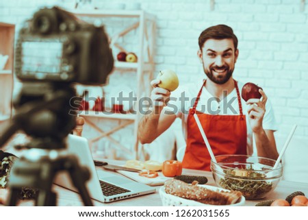 Blogger Makes a Video. Blogger is Smiling Beard Man. Video About a Cooking. Camera Shoots a Video. Laptop and Different Food on Table. Man Showinf an Apples. Man in Studio Interior.