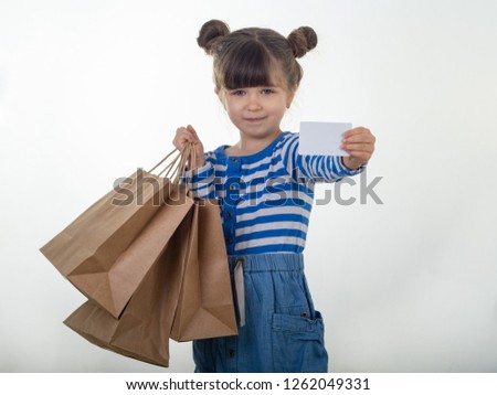 Happy child holding discount white card  and shopping bags in her hands. Kid with credit card. Little girl showing empty blank paper note copy space. White background.