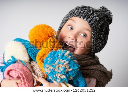 surprised excited little girl wearing knitted hat, scarf and sweater, holding a pile of hats, Child on light background. Fashion, choice, sale concept.