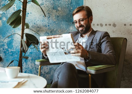 Smiling businessman wearing suit reading newspaper while sitting at the cafe and drinking coffee