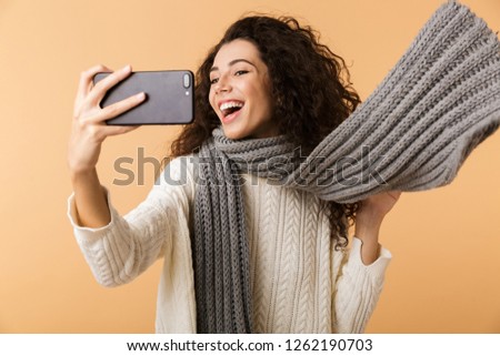 Cheerful young woman wearing winter scarf standing isolated over beige background, taking a selfie