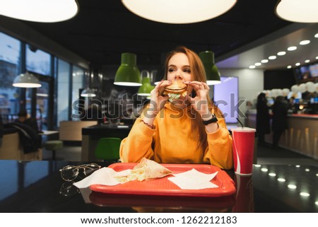 Girl bites the burger and gets pleasure. Fast Food Concept. Portrait of a beautiful girl in a orange hoodie bites an appetizing burger against the background of a fast-food restaurant.