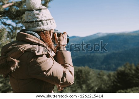 
brunette girl in wool hat and brown coat takes a picture in the middle of the mountain
