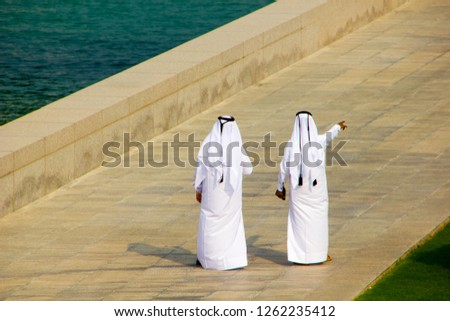 Two Qatari business men wearing white thob are meeting outside on a public place and one is pointing to a certain direction. Typical Arab fashion Royalty-Free Stock Photo #1262235412