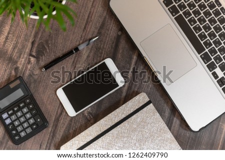 open laptop, phone with black screen, black calculator, closed silver notebook, green plant and black pen on ructic table