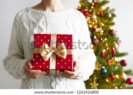 Cropped shot of unrecognizable young woman wearing white knitted sweater, holding a present with festive red wrapping, tied with golden bow, christmas tree on background. Close up, copy space.