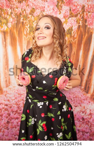 beautiful young girl standing in black pin up dress and having fun playing with glazed pink donuts