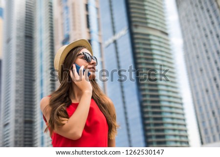 Portrait of young woman in red dress, sunglasses and summer hat talking on the phone on downtown skycrapers background
