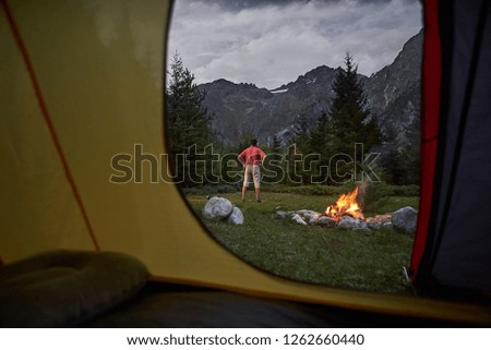 View from inside tent at sunset. Alone tourist, hiker standing at bonfire