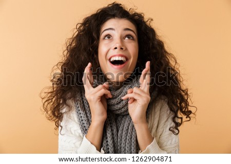 Cheerful young woman wearing winter scarf standing isolated over beige background, holding fingers crossed for good luck