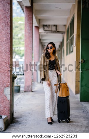 A full-body portrait of a young, Indian Asian (Pubjabi) woman standing on the street with her luggage. She looks elegant, wealthy and stylish with her pant suit outfit and sunglasses.