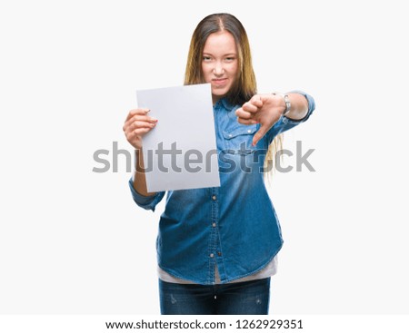 Young caucasian woman holding blank paper sheet over isolated background with angry face, negative sign showing dislike with thumbs down, rejection concept