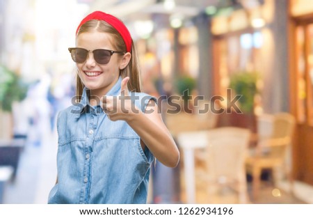 Young beautiful girl wearing sunglasses over isolated background doing happy thumbs up gesture with hand. Approving expression looking at the camera with showing success.