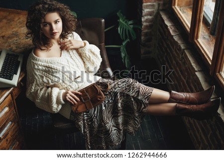 Full body fashion portrait of young beautiful confident woman wearing cozy white knitted sweater, animal print skirt, cowboy style boots, holding small brown bag, posing at loft interior Royalty-Free Stock Photo #1262944666