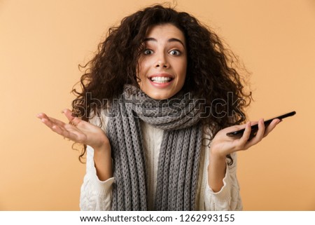 Smiling confused young woman wearing sweater and scarf holding mobile phone isolated over beige background