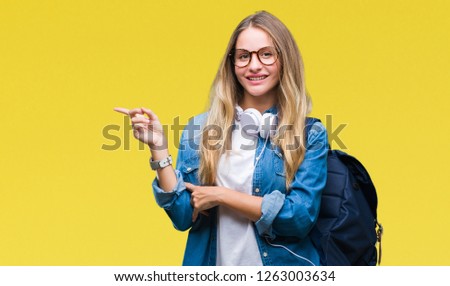 Young beautiful blonde student woman wearing headphones and glasses over isolated background with a big smile on face, pointing with hand and finger to the side looking at the camera.
