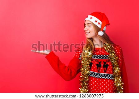 Beautiful girl in a red sweater and Santa Claus hat, with tinsel on her neck shows her finger on an empty space on a red background