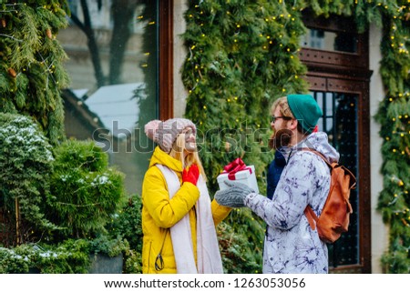 Picture showing young hipster couple in colorful bright outwear with present in the street in snowfall winter day. Emotions and give a gift concept